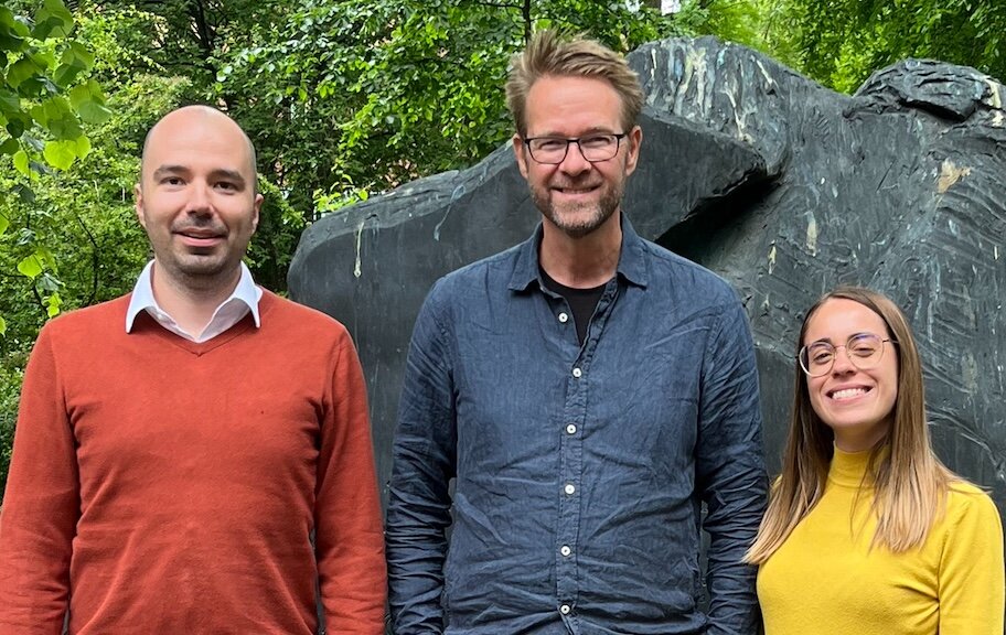 The three researchers stand side by side smiling in front of a statue