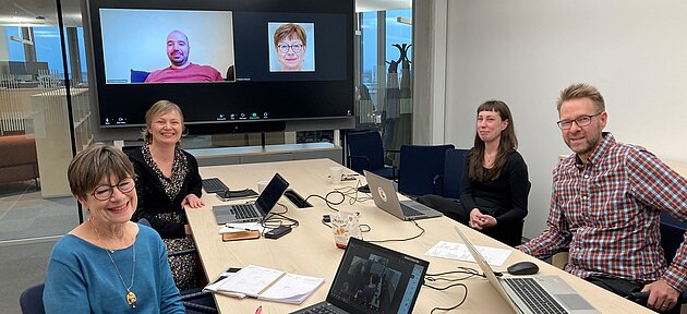 Researchers sitting around a table. One person is participating in Zoom on a big screen at the back of the room.