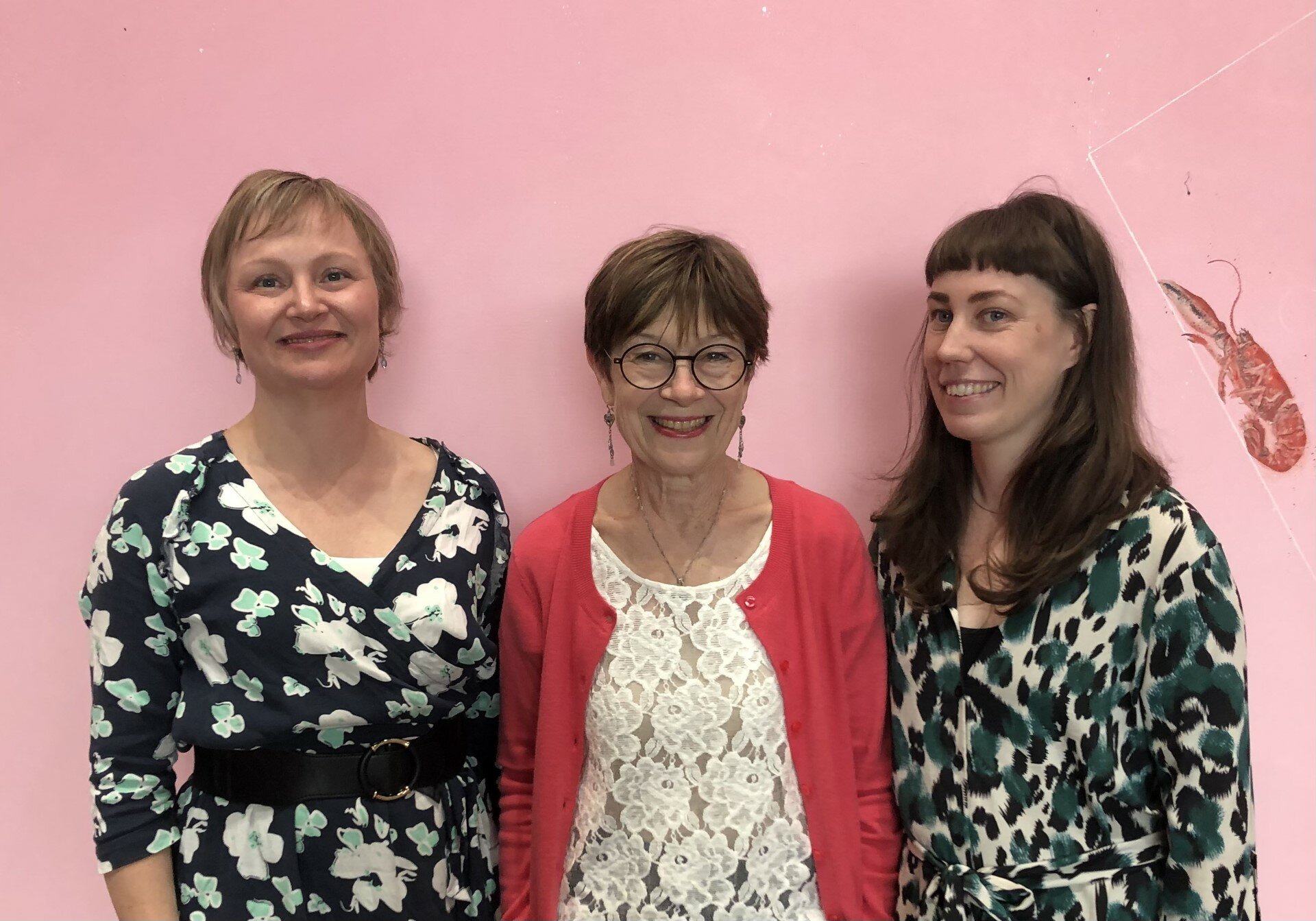 Three people are standing in front of a pink background. They smile at the camera. To the right of them, there is a cooked ocean crayfish