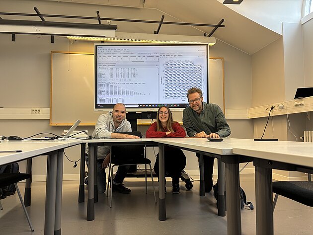 The P2 team in front of a screen with data. The three project members are smiling into the camera .