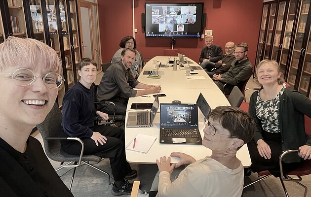 A room full of people seated at a long table. At the end of the table, a screen on the wall. The screen shows participants in a Zoom room.