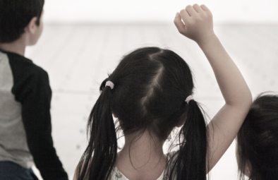 A group of children seen from behind. One child raises their hand.