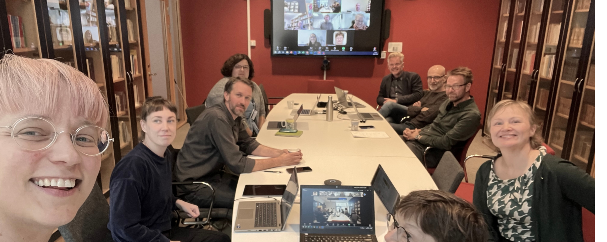 A seminar room full of people, even more are displayed on a screen of a zoom room on the back wall of the room.