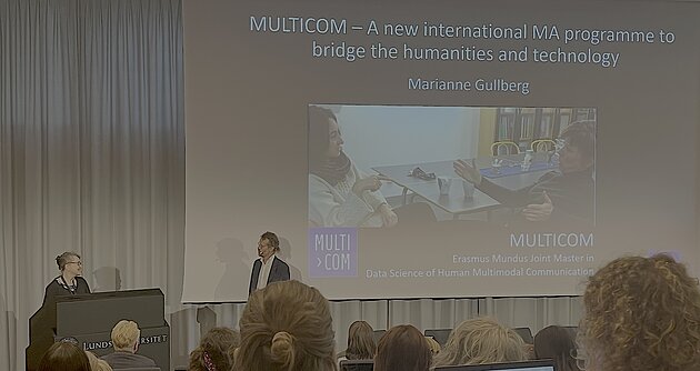 Two people are placed in front of a big screen projecting the title slide of a powerpoint presentation. The people are looking at eachother and smiling. The title slide reads "MULTICOM – A new international MA programme to bridge the humanities and technology"