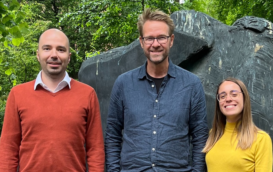 The three researchers stand side by side smiling in front of a statue